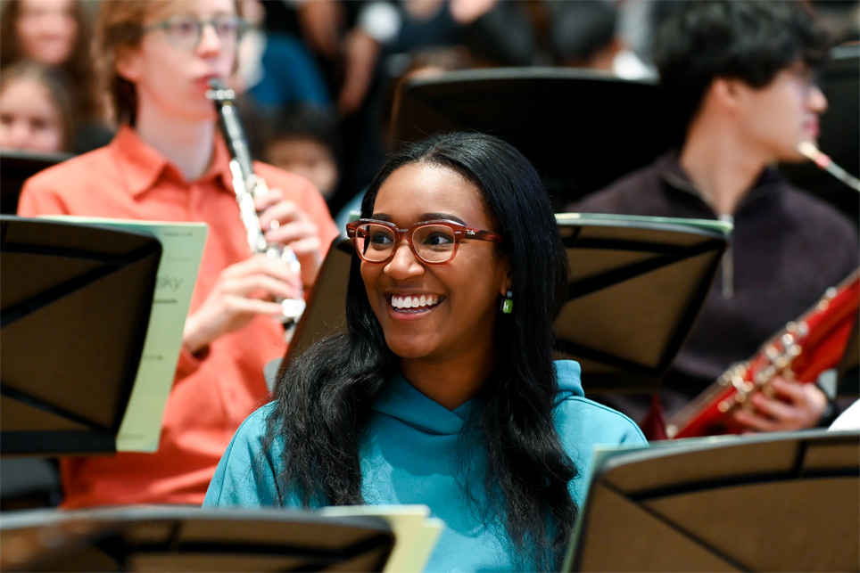 A black student wearing a blue top, holding her flute, smiling in the woodwind section in a orchestra rehearsal.